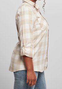 Donna che indossa una camicia a maniche lunghe beige e bianca a scacchi con le maniche arrotolate, abbinata a jeans blu, su uno sfondo neutro.