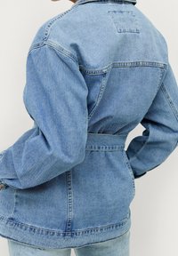 Ljusblå denimjacka med bälte i midjan, långa ärmar och ryggsöm. Utmärkande sömmar och en patchdetalj på övre delen av ryggen.