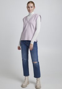 Top sin mangas en color lila con escote en V, superpuesto a un cuello de tortuga blanco. Llevado con jeans azules desgastados y botines beige.