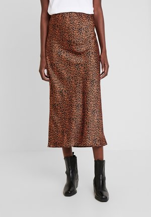 Kobieta w brązowo-czarnej spódnicy midi w cętki, białej bluzce i czarnych skórzanych botkach, stojąca na jasnoszarym tle.