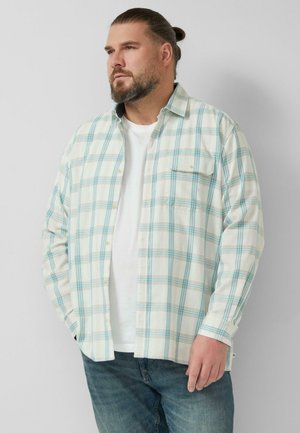 Brodaty mężczyzna z włosami związanymi do tyłu, ubrany w biały t-shirt i jasno-niebieską koszulę w kratę, stoi z rękami w kieszeniach na tle białej ściany.