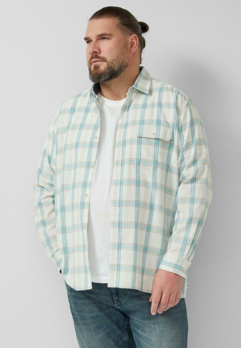 Bärtiger Mann mit zurückgebundenem Haar, trägt ein weißes T-Shirt und ein hellblau karierte Hemd, steht mit den Händen in den Taschen vor einer weißen Wand.