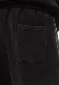 Čierne športové nohavice s textúrovanou tkaninou, obsahujúce vrecko na zadnej strane. Vrecko má zaoblené rohy a je našité čiernou niťou.