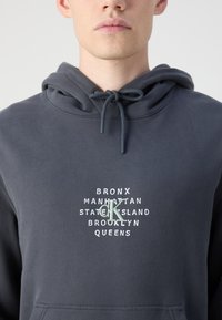 Tmavě šedá mikina s kapucí a stahovací šňůrkou, s bílými a zelenými textovými grafikami názvů čtvrtí a centrálním logem "CK". Měkká strukturovaná tkanina.
