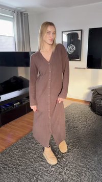 Γυναίκα με ξανθά μαλλιά που φοράει μακρύ καφέ φόρεμα με κουμπιά και μπεζ μπότες, στέκεται πάνω σε γκρι χαλί σε ένα σαλόνι.