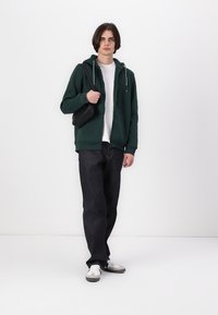 Zöld kapucnis pulóver, fehér póló, sötét farmernadrág és szürke sportcipő. A modellen egy fekete válltáska van. Egyszerű design, laza fazon.