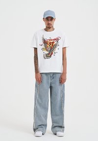 Bílý grafický tričko s červeným a žlutým designem orla, doplněné o zničené světle modré široké džíny a bílé tenisky. Světle modrá kšiltovka.