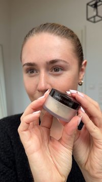 Gjennomskinnelig krukke med svart lokk, merket "LAURA MERCIER." Inneholder pudder med en glatt tekstur. Hender som holder krukken, nøytral bakgrunn.