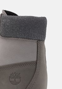 Botă din piele gri, cu un guler moale din fetru gri, corp texturat și detalii de cusătură. Include un logo cu un copac embosat pe lateral.