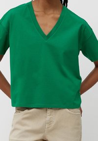 Persona con una camiseta verde de cuello en V y mangas cortas, combinada con pantalones beige, con las manos reposando detrás de la espalda.