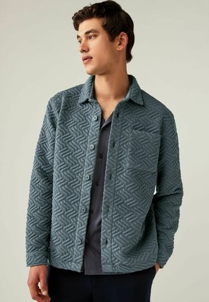 Jachetă texturată cu buton în nuanță de teal estompat, având un model în zigzag, guler, buzunar frontal și o cămașă închisă, în contrast, dedesubt.