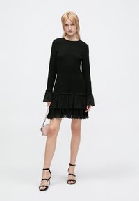 Vestido negro con mangas largas, cuerpo texturizado y falda con volantes en capas. Mangas acampanadas transparentes y detalle de encaje. La modelo sostiene un pequeño bolso rosa.