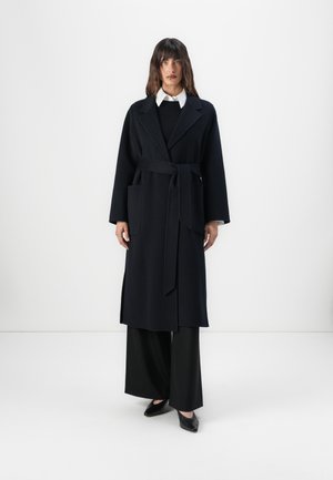 Cappotto lungo in lana nera con vita accentuata da una cintura, collo a rever e tasche laterali, abbinato a pantaloni neri a gamba larga e scarpe appuntite.