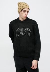 Pulover negru tricotat cu "OBEY" brodat în alb, asortat cu o căciulă bej cu model cu buline.