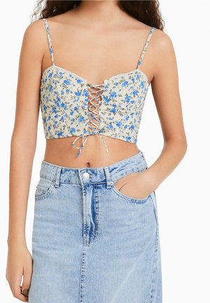 Femme portant un crop top blanc à fleurs bleues avec un laçage sur le devant, associé à une jupe en jean bleu clair délavée.