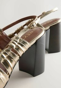 Gold strappy high-heeled sandals with thick black block heels and adjustable buckle straps on a light background.