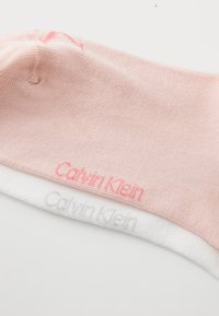 Šviesiai rožinė kojinė iš elastingos medžiagos, su „Calvin Klein“ logotipu rožine ir pilka spalvomis ant baltos apvado. Minkšta tekstūra, glotnus dizainas.