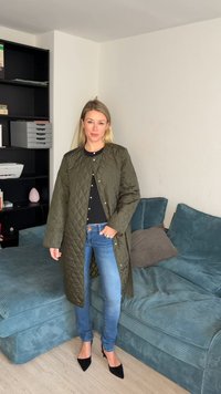 Γυναίκα στέκεται μέσα σε χώρο φορώντας ένα μακριά μπουφάν καπιτονέ σε ελιά, μαύρο τοπ, μπλε τζιν και μαύρες γόβες κοντά σε μια βεραμάν καναπέ και ραφιέρα.