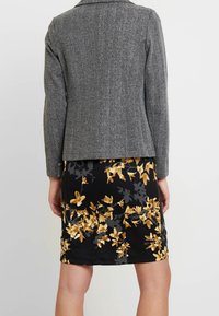 Sivý houndstooth blazer s přizpůsobeným střihem, zkombinovaný s černou sukní se žlutými květinovými vzory. Texturované látky a strukturovaný design.