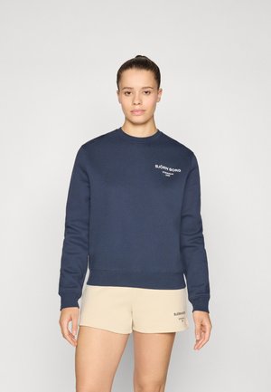 Mørkeblå hettegenser med rund hals, lange ermer og ribbede mansjetter; har et hvitt tekstlogo. Kombinert med beige shorts.