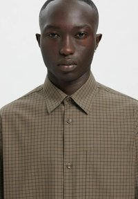 Brązowa koszula w kratę z zapinanym na guziki przodem i spiczastym kołnierzykiem. Posiada kieszeń na wysokości klatki piersiowej oraz fakturowaną tkaninę z delikatnymi poziomymi i pionowymi liniami.