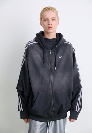 Oversized sort zip-up hættetrøje med grå gradient, store forlommer og hvide tre-stribede accenter på ærmerne. Der er et logo på forsiden.