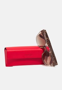 Lunettes de soleil teintées marron avec des montures en écaille reposant contre un étui rouge texturé de la marque GUESS.