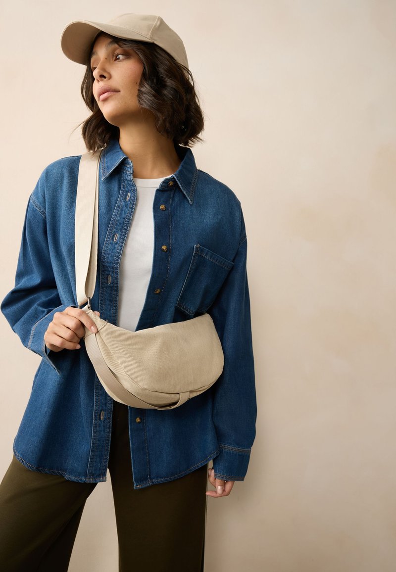 Beige Umhängetasche aus weichem Stoff, mit einer halbmondförmigen Silhouette und dezenter gerippter Textur. Getragen über einem Jeanshemd.