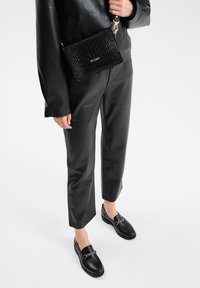 Zwarte leren loafers met een kettingaccent, gecombineerd met zwarte nepleer broek en een gestructureerde zwarte crossbody tas, wat zorgt voor een monochrome look.