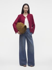 Femme en veste rouge texturée, top blanc, jean bleu large et sandales noires tenant un grand sac en daim marron sur fond blanc.