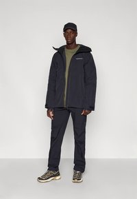 Giacca nera con cappuccio e zip frontale, abbinata a pantaloni neri e una maglietta verde sotto. Caratteristiche includono una texture liscia e una grafica minimale.