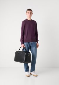 Borsa da viaggio in pelle nera con forma arrotondata, doppio manico e dettagli in metallo argento. Indossata da una persona in un maglione viola e jeans.