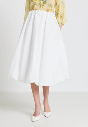 Jupe midi blanche associée à des talons blancs pointus, modèle portant un chemisier jaune à fleurs avec des manches trois-quarts.