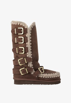 Botte en suède marron hauteur genou avec des accents tressés, cinq sangles à boucle et un bord supérieur légèrement scallopé. Semelle texturée pour une meilleure adhérence.