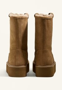 Brown suede ankle boots with a plush lining, featuring a flat platform sole and dual loop pull tabs at the top for easy wear.