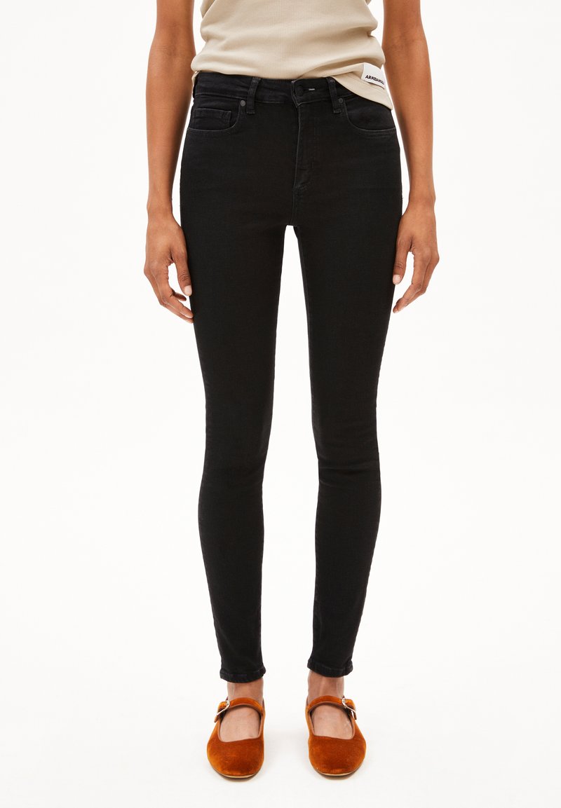 Jeans skinny en denim noir avec une texture lisse, taille haute et design à cinq poches. Associés à des pantoufles orange.