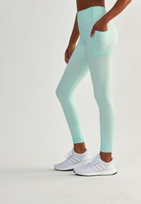 Miętowe legginsy mają wysoki pas i kieszeń z boku. Materiał wydaje się elastyczny, zestawione z białymi butami sportowymi.