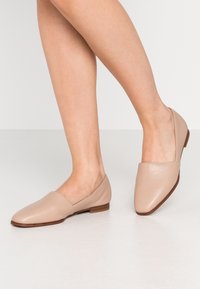 Chaussures à enfiler en cuir nude, à bout pointu et talon carré bas, présentant une texture lisse et un design minimal.