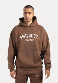 Sweat-shirt à capuche marron en tissu doux, doté d'une poche frontale, de poignets côtelés et de l'inscription "SMILODOX ATHL. WEAR" sur la poitrine.