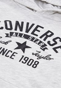 Šedá mikina s výrazným čiernym textom "CONVERSE ALL STAR", grafickým prvkom hviezdy a nápisom "Taylor" písaným kurzívou. Mäkká textúra s klasickým dizajnom.