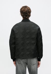 Άνδρας με κυματιστά καστανά μαλλιά φοράει μαύρο καπιτονέ μπουφάν και ξεβάμμένο γκρι τζιν, στέκεται με την πλάτη προς τα εμπρός σε μονόχρωμο λευκό φόντο.