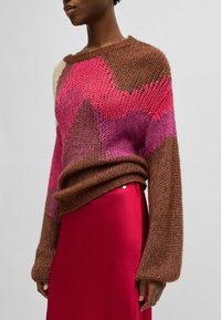 Brown and pink knitted sweater with wide sleeves, featuring a geometric pattern; paired with a smooth red skirt. Textured, loose fit.