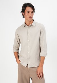 Camisa botão bege clara com um padrão texturizado, colarinho abotoado e mangas arregaçadas, usada com calças listradas em castanho claro.