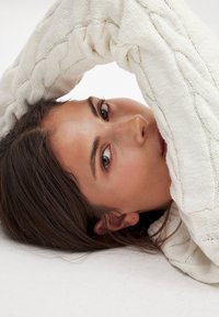 Model mit langen braunen Haaren, die auf einer weißen Oberfläche ausruht, teilweise bedeckt mit einem strukturierten, cremefarbenen Zopfmuster-Pullover.