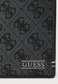 Musta lompakko, jossa on kuvioitu kangaspinta ja kohokuvioitu logokuvio. Hopeinen "GUESS"-logolevy ja kontrastisommitelut lisäävät yksityiskohtia.