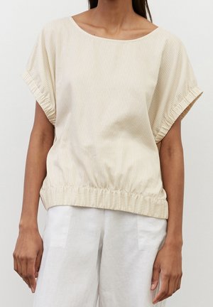 Vrouw draagt een beige blouse met korte mouwen met subtiele verticale strepen en een elastische zoom, gecombineerd met losse witte broek.