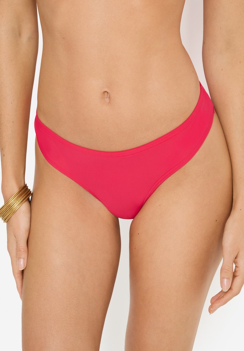 Moteris dėvi ryškiai rožinius bikini apatinius ir auksinius apyrankes ant kairės riešo, matoma nuo liemens vidurio iki viršutinės šlaunų dalies, baltame fone.