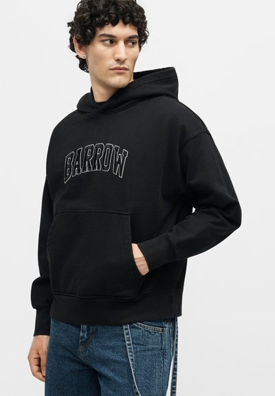 Άνδρας με σγουρά μαλλιά που φοράει μαύρο φούτερ "BARROW" και μπλε τζιν, με το ένα χέρι στην τσέπη, κοιτάζοντας προς τα αριστερά.