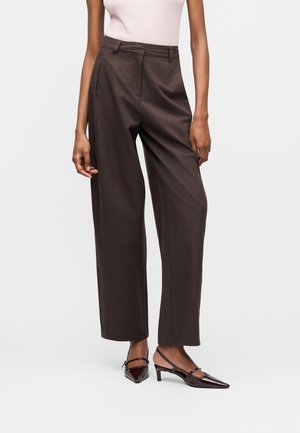 Žena nosiaci tmavohnedé nohavice s vysokým pásom a širokými nohavicami v kombinácii s bordovými špicatými slingback lodičkami, stojaca pred jednoduchým pozadím.