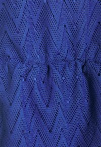 Modrá pletená látka se vzorem zigzag chevron a malými dírkami, mírně nařasená ve středu.